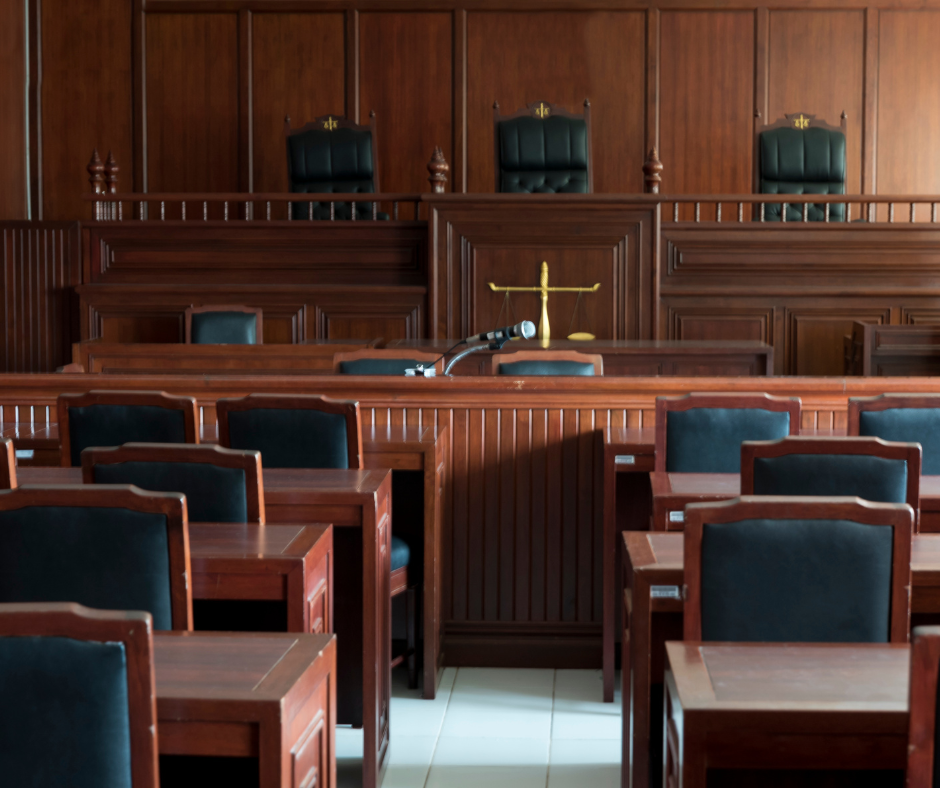 Image depicts an empty courtroom with a row of chairs and a judge's table.