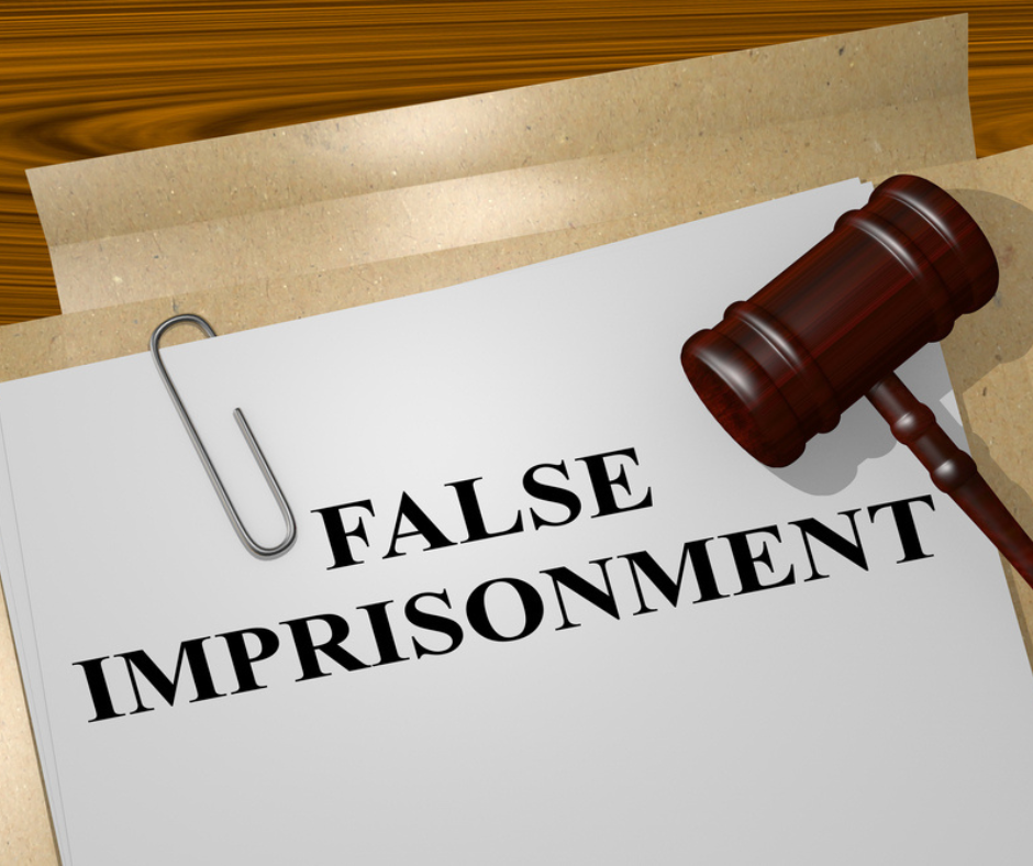 Piece of white paper that says false imprisonment in all black capital letters. The piece of white paper has a paper clip on it and is rested on beige pieces of paper. There is a brown gavel laying on the white piece of paper. 